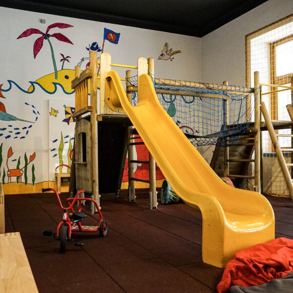 Innenspielzimmer mit einer gelben Rutsche, einem hölzernen Klettergerüst, Bänken, einem roten Dreirad und farbenfrohen Wandmalereien mit Meeresbewohnern und Pflanzen.