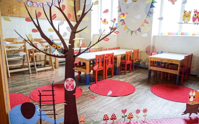 Ein hell dekoriertes Klassenzimmer mit kleinen Tischen, Stühlen, roten Teppichen und verspielten Wand- und Fensterbildern von Bäumen, Eulen und einer Elefantenleiter.