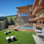 Ein modernes Holzhotel mit Balkonen und Blick auf einen grasbewachsenen Hof mit Liegestühlen und einem kleinen Pool, umgeben von Bäumen und fernen Hügeln unter einem klaren blauen Himmel.