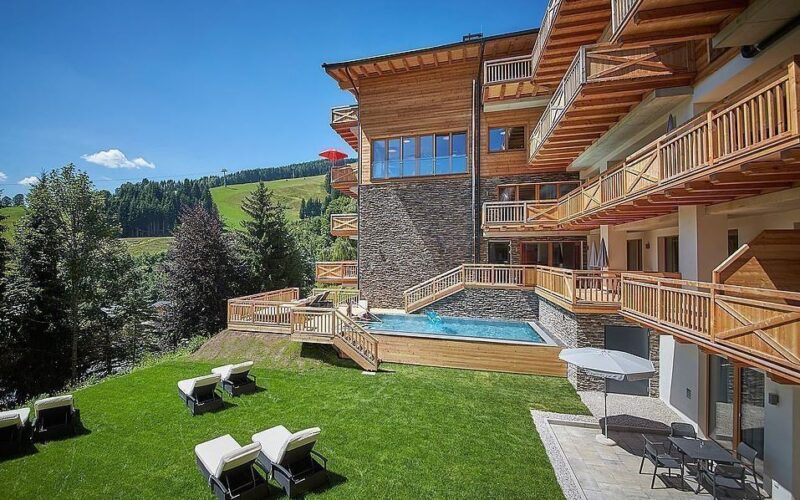 Ein modernes Holzhotel mit Balkonen und Blick auf einen grasbewachsenen Hof mit Liegestühlen und einem kleinen Pool, umgeben von Bäumen und fernen Hügeln unter einem klaren blauen Himmel.