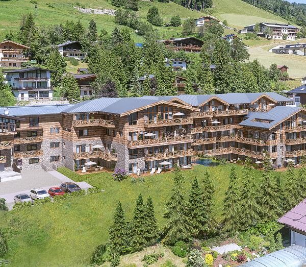 Ein großes, hölzernes Gebäude im Chalet-Stil mit Balkonen steht auf einem Hügel, umgeben von Gras, Bäumen und anderen ähnlichen Häusern in einer bergigen Gegend.