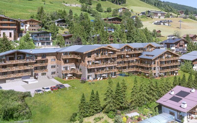 Ein großes, hölzernes Gebäude im Chalet-Stil mit Balkonen steht auf einem Hügel, umgeben von Gras, Bäumen und anderen ähnlichen Häusern in einer bergigen Gegend.