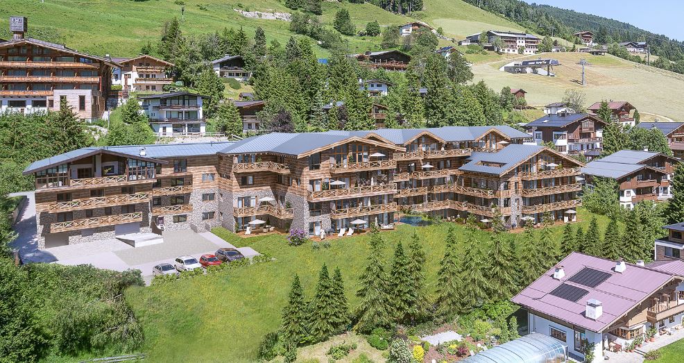 Ein großes, hölzernes Gebäude im Chalet-Stil mit Balkonen steht auf einem Hügel, umgeben von Gras, Bäumen und anderen ähnlichen Häusern in einer bergigen Gegend.