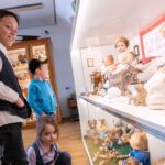Drei Kinder betrachten eine Vitrine mit alten Puppen und Teddybären in einer Museumsumgebung.