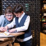 Ein Erwachsener und zwei Kinder arbeiten an einem Tisch in einem Museum oder Lernzentrum an einer pädagogischen Aktivität, mit Ausstellungsregalen im Hintergrund.