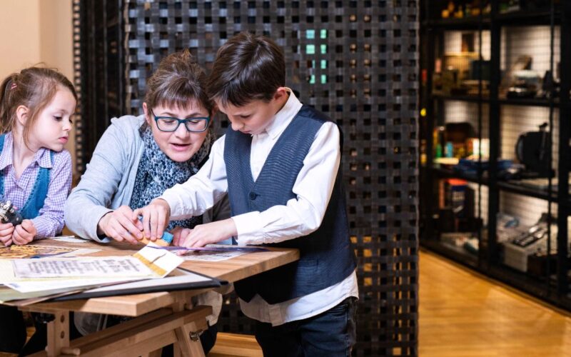 Ein Erwachsener und zwei Kinder arbeiten an einem Tisch in einem Museum oder Lernzentrum an einer pädagogischen Aktivität, mit Ausstellungsregalen im Hintergrund.