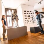 Zwei Personen betrachten und interagieren mit mittelalterlichen Rüstungen und Waffen in einem Ausstellungsraum mit Holzboden und weißen Wänden.