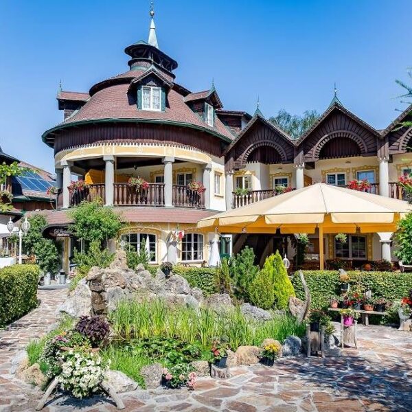 Ein großes, verziertes Gebäude mit Holzakzenten und Balkonen, umgeben von üppigen Gärten, einer Steinterrasse, einem Teich und Sitzgelegenheiten im Freien unter einem gelben Sonnenschirm.