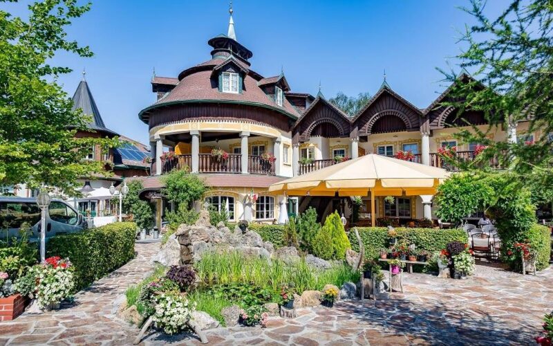 Ein großes, verziertes Gebäude mit Holzakzenten und Balkonen, umgeben von üppigen Gärten, einer Steinterrasse, einem Teich und Sitzgelegenheiten im Freien unter einem gelben Sonnenschirm.