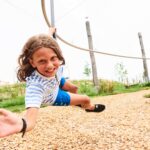 Ein Kind in einem gestreiften Hemd und blauen Shorts fährt auf einer Seilrutsche im Freien, lächelt und streckt eine Hand nach vorne, während Holzspäne auf dem Boden liegen.