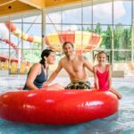 Ein Mann und zwei Kinder sitzen auf einem großen roten aufblasbaren Ring in einem Indoor-Wasserpark, mit Wasserrutschen und großen Fenstern im Hintergrund.