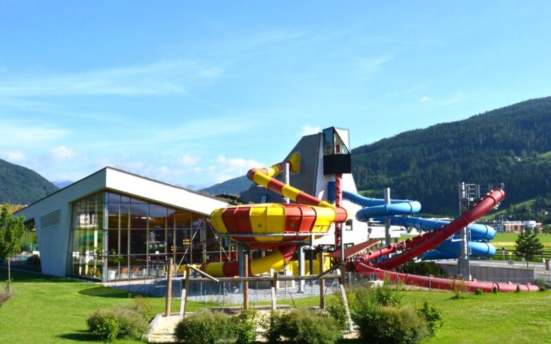 Ein modernes Wasserparkgebäude mit großen Glasfenstern und mehreren farbenfrohen Wasserrutschen im Freien vor einer Kulisse aus grünen Hügeln und blauem Himmel.