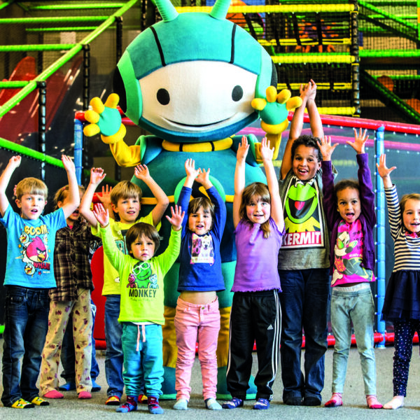 Neun Kinder stehen mit erhobenen Armen vor einem großen, bunten Maskottchen in einem Indoor-Spielbereich mit Klettergerüsten und Netzen.