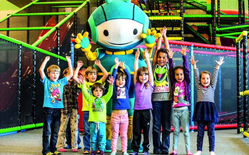 Neun Kinder stehen mit erhobenen Armen vor einem großen, bunten Maskottchen in einem Indoor-Spielbereich mit Klettergerüsten und Netzen.