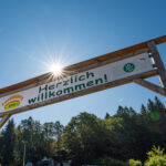 Ein großes Holzschild mit der Aufschrift „Herzlich willkommen!“ steht vor einem klaren blauen Himmel mit Sonnenlicht und Bäumen im Hintergrund.
