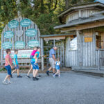 Ein Mann und fünf Kinder gehen in einem bewaldeten Außenbereich auf ein rustikales hölzernes Eingangstor mit Informationsschildern zu.