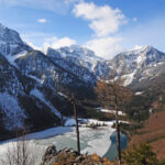 Ein teilweise zugefrorener See, umgeben von schneebedeckten Bergen und bewaldeten Hängen unter einem blauen Himmel mit vereinzelten Wolken.