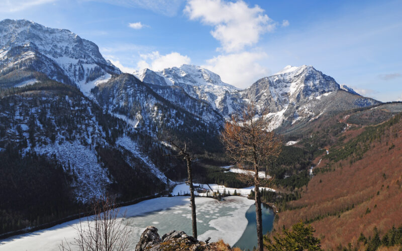 Ein teilweise zugefrorener See, umgeben von schneebedeckten Bergen und bewaldeten Hängen unter einem blauen Himmel mit vereinzelten Wolken.