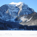 Schneebedeckter Berg mit bewaldetem Fuß und einem Haus im Vordergrund unter einem klaren blauen Himmel.