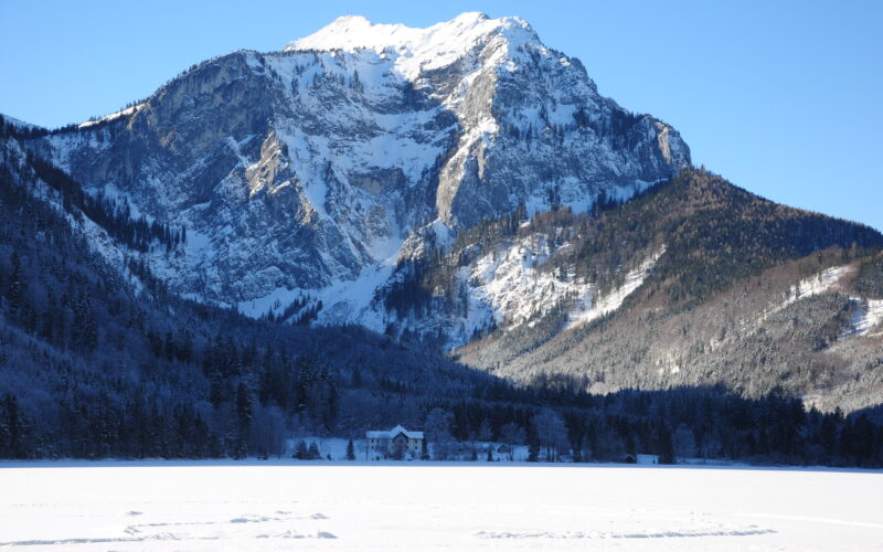 Schneebedeckter Berg mit bewaldetem Fuß und einem Haus im Vordergrund unter einem klaren blauen Himmel.