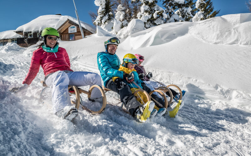 Vier Personen, zwei Erwachsene und zwei Kinder, sitzen auf Holzschlitten im Schnee, tragen Helme und Winterkleidung, im Hintergrund verschneite Bäume und eine Hütte.