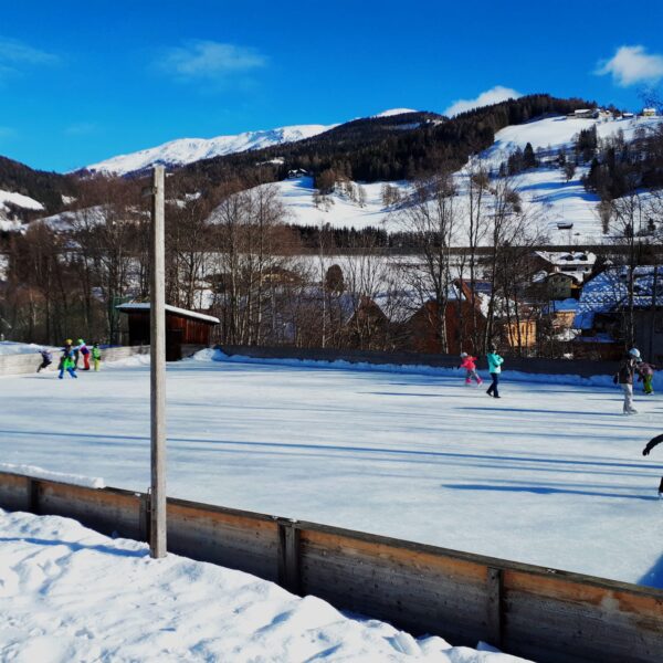Menschen beim Schlittschuhlaufen auf einer Freiluft-Eisbahn, umgeben von Schnee, Bäumen und Bergen unter blauem Himmel.