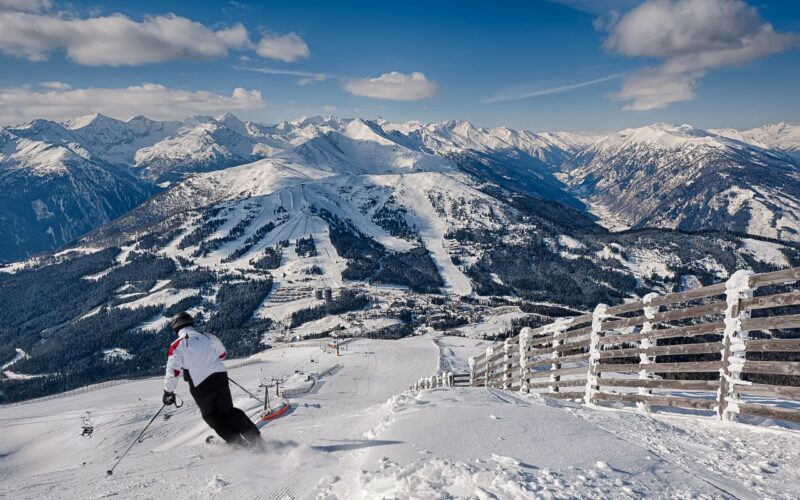 Ein Skifahrer in einer weißen Jacke fährt auf einer verschneiten Bergpiste bergab, mit einem Holzzaun auf der rechten Seite und schneebedeckten Gipfeln im Hintergrund unter einem teilweise bewölkten Himmel.