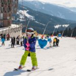 Ein Kind in Skiausrüstung hält ein Schleppseil auf einer verschneiten Piste, im Hintergrund sind andere Skifahrer und ein Gebäude zu sehen, in der Ferne die Berge.
