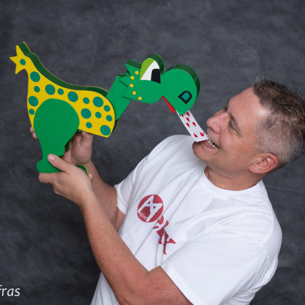 Ein Mann in einem weißen T-Shirt hält eine grün-gelb gepunktete Dinosaurierpuppe, die in der Nähe seines Mundes auf einer Spielkarte herumzukauen scheint.