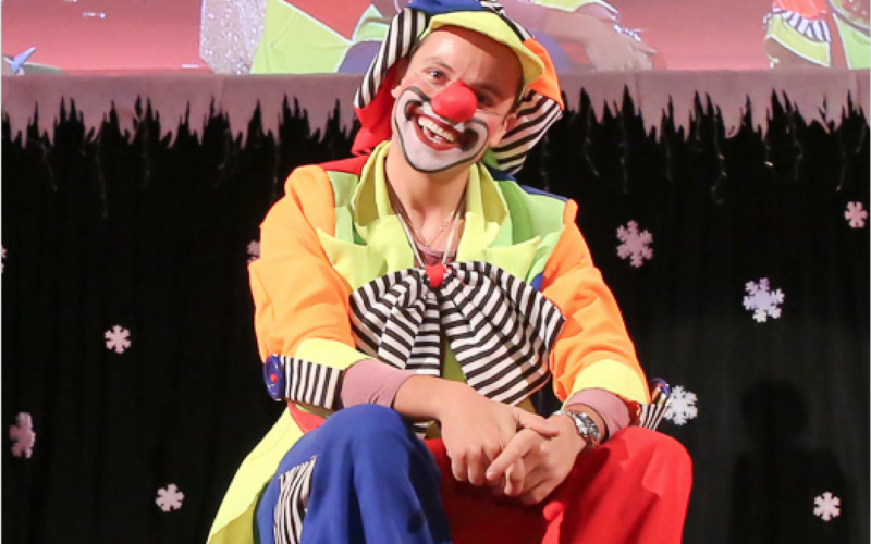 Eine als Clown verkleidete Person mit roter Nase, buntem Kostüm und Gesichtsbemalung sitzt auf der Bühne vor einem mit Schneeflocken dekorierten Hintergrund.