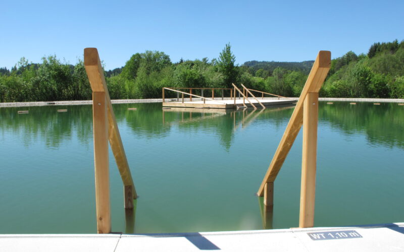 Ein Naturschwimmbad mit Holzgeländer und einer kleinen Holzplattform auf dem Wasser, umgeben von Grün unter einem klaren blauen Himmel.