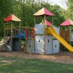 Spielplatz im Freien mit Rutschen, Kletterwänden und Brücken auf einer mit Mulch bedeckten Fläche, umgeben von Gras und Bäumen.