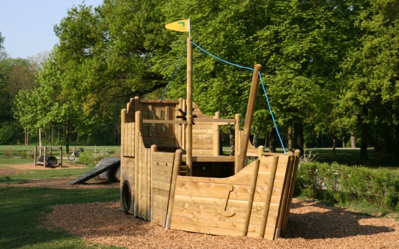 Ein hölzernes Spielgerät in Form eines Schiffes steht auf Mulch in einem Park, mit Bäumen und Gras im Hintergrund.