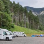 Drei Wohnmobile stehen auf einem Schotterplatz neben mehreren Zelten, vor der Kulisse hoher Kiefern und Berge unter einem wolkenverhangenen Himmel.