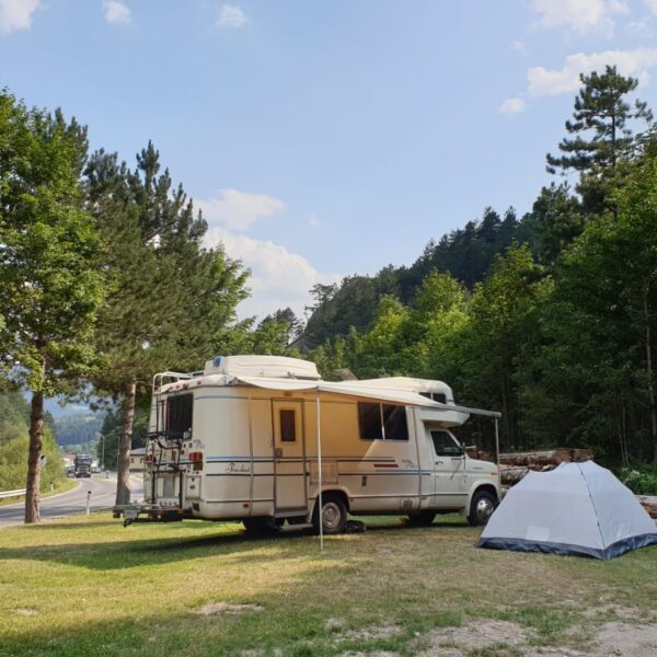 Ein weißes Wohnmobil und ein graues Zelt stehen auf einer Wiese in der Nähe einer Straße, umgeben von Bäumen und Hügeln unter einem teilweise bewölkten Himmel.