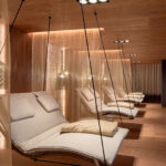 Moderne Entspannungslounge mit gepolsterten Schaukelstühlen, gedämpfter Beleuchtung, Holzböden und -decken sowie halbtransparenten Vorhängen, die jeden Sitzbereich abtrennen.