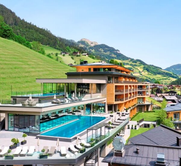 Modernes, mehrstöckiges Resort mit Außenpools und Lounge-Bereichen, das auf einem Hügel mit grünen Hängen und Bergen im Hintergrund unter einem klaren blauen Himmel liegt.