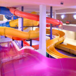 Farbenfrohe Indoor-Wasserrutschen mit gelben, rosa, roten und blauen Röhren, die sich um weiße Säulen in einer gut beleuchteten Freizeitanlage winden.