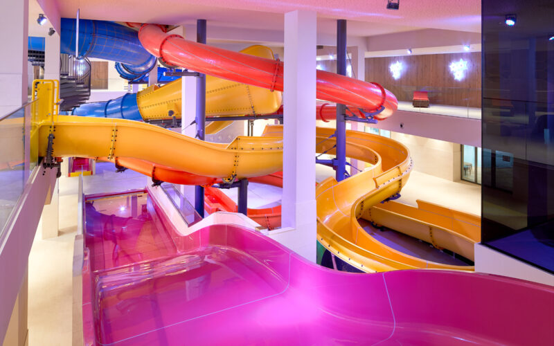 Farbenfrohe Indoor-Wasserrutschen mit gelben, rosa, roten und blauen Röhren, die sich um weiße Säulen in einer gut beleuchteten Freizeitanlage winden.