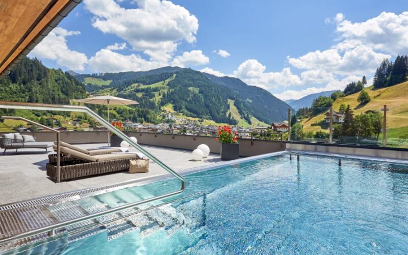 Außenschwimmbad auf einer Terrasse mit Liegestühlen und Blick auf die Berge unter einem teilweise bewölkten Himmel.