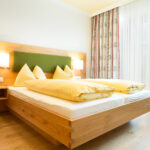 Ein modernes Hotelzimmer mit einem hölzernen Doppelbett, einem grünen Kopfteil, gelber Bettwäsche, zwei Wandlampen, einem Beistelltisch mit einem Telefon und einem Fenster mit durchsichtigen und geblümten Vorhängen.