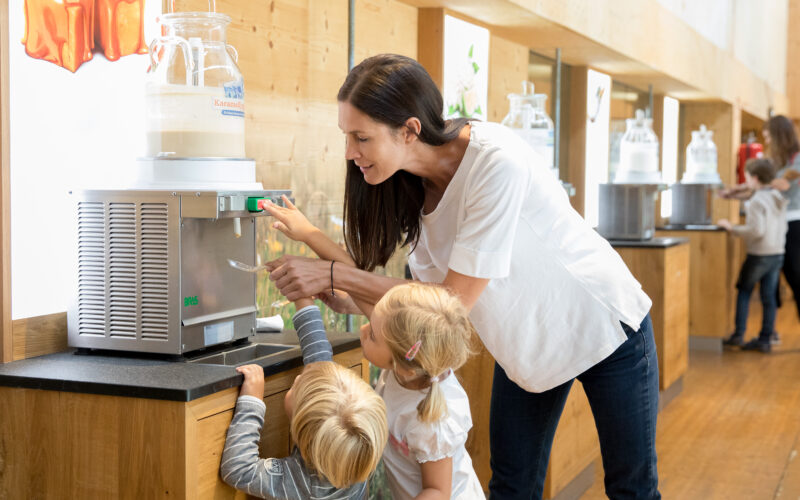 Eine Frau hilft zwei kleinen Kindern, einen Milchspender in einem hellen Innenraum mit Holzböden und Displays zu bedienen.