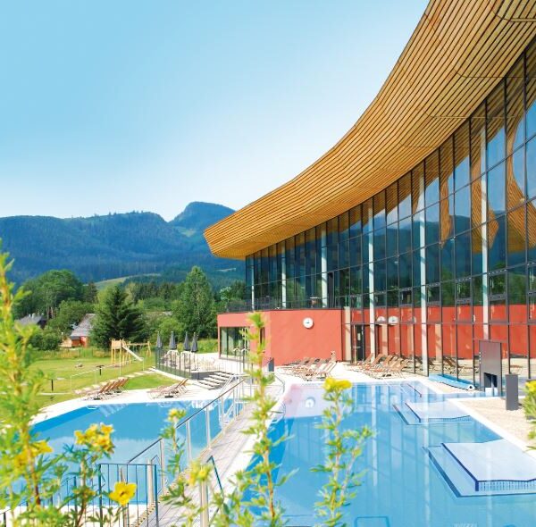 Modernes Gebäude mit großen Glasfenstern neben Außenpools, umgeben von grünen Rasenflächen und fernen Bergen unter einem klaren blauen Himmel.