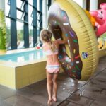 Ein Kind im Badeanzug steht drinnen an einem kleinen Pool und hält einen großen aufblasbaren Donut-Schwimmer mit Streuseln und einen rosa Flamingo-Schwimmer dahinter.