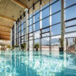 Hallenbad mit klarem Wasser, große, raumhohe Fenster mit Blick auf die schneebedeckten Berge und Holzdeckenpaneele. Topfpflanzen säumen die Fenster.