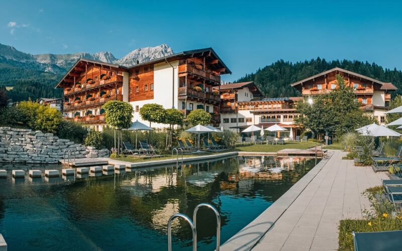 Ein Hotel mit Holzbalkonen liegt an einem angelegten Naturschwimmteich, umgeben von Liegestühlen, Sonnenschirmen und den Bergen im Hintergrund.