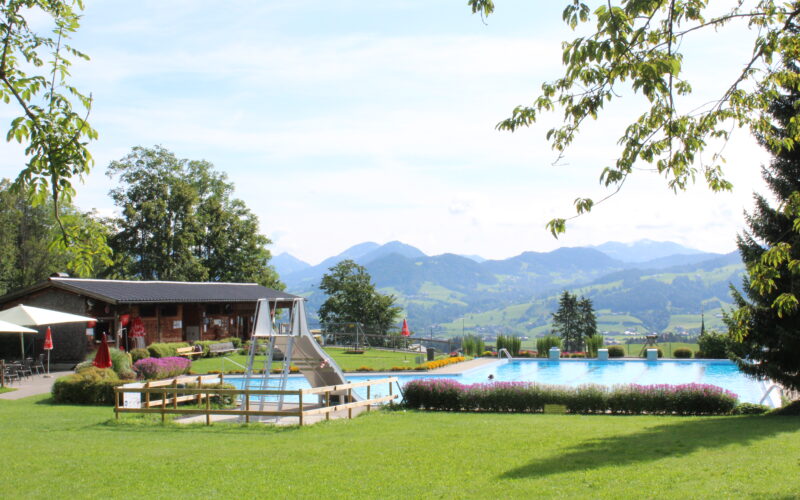 Außenschwimmbad mit einem Holzgebäude in der Nähe, umgeben von grünem Gras und Bäumen, mit Bergen im Hintergrund unter einem teilweise bewölkten Himmel.