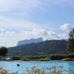 Außenschwimmbad mit einer Person im Wasser, umgeben von Grün und Bergen unter einem blauen Himmel mit vereinzelten Wolken.