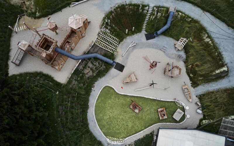 Luftaufnahme eines Spielplatzes mit Holzkonstruktionen, Rutschen, Klettergerüsten, Kieswegen und einem kleinen grünen Teich, der von Gras und Vegetation umgeben ist.