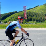 Ein Radfahrer in blauem Trikot und Helm fährt mit seinem Rennrad auf einer asphaltierten Straße, im Hintergrund sind grasbewachsene Hügel, Bäume und Flaggen unter einem klaren blauen Himmel zu sehen.